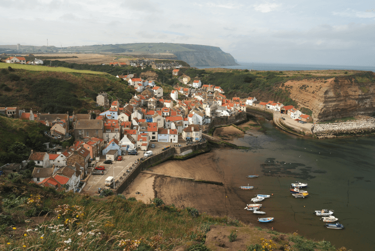 Staithes, one of North Yorkshire's prettiest villages, is a cluster of white houses with red roofs at the back of a deeply-sided cove on the coast. Boats float in the bay out front, and in the background is another headland jutting into the sea.